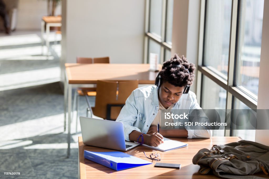 Teenage boy studies in school library African American teenage boy writes something in a notebook while studying in the campus library. An open laptop is on the table. He is wearing wireless headphones. Student Stock Photo