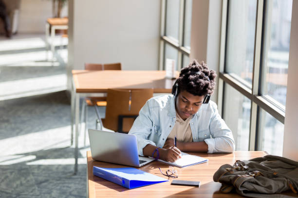 Teenage boy studies in school library African American teenage boy writes something in a notebook while studying in the campus library. An open laptop is on the table. He is wearing wireless headphones. education building photos stock pictures, royalty-free photos & images
