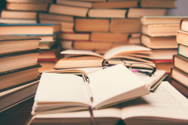 istock Education learning concept with open book and notebook on the table. Stack piles of books on reading desk and glasses in bookshelves background 1137626325