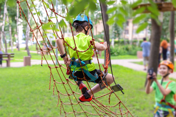 little boy in safety equipment climbing on rope wall at adventure park. fiend making a photo shot on smartphone. children summer sport extreme outdoor activity. back view - tree skill nature horizontal imagens e fotografias de stock