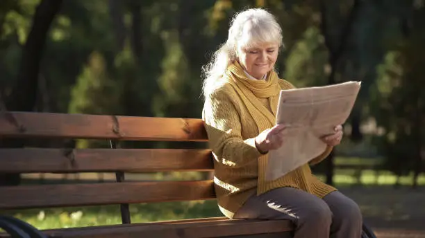 Concentrated mature woman reading newspaper sitting on bench in park, retirement