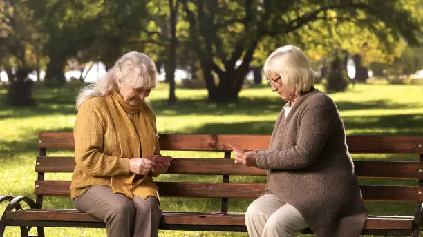 Couple of adult ladies playing cards sitting on bench in park, leisure activity