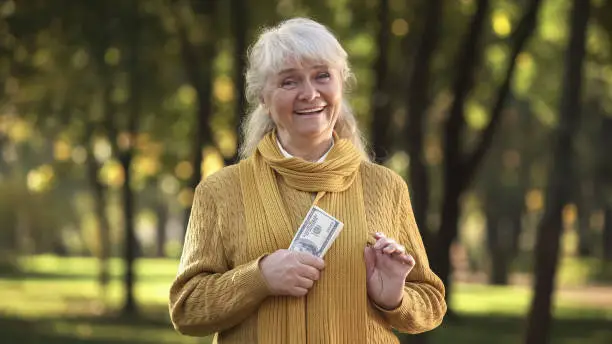 Energetic old lady holding stack of dollar bills in park, planned retirement
