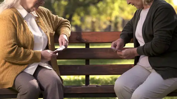 Two old women enjoying cards playing on bench in park, elderly friends leisure