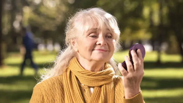Mature woman looking at her reflection in compact mirror sitting in park