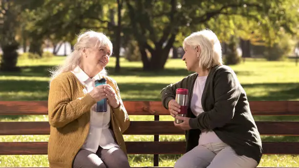 Two old ladies drinking hot tea from travel mugs, sitting on bench in sunny park