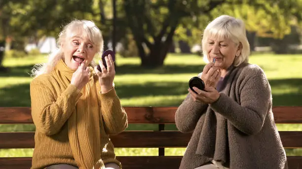 Two old ladies doing make up, sitting on bench in park, anti-age cosmetics