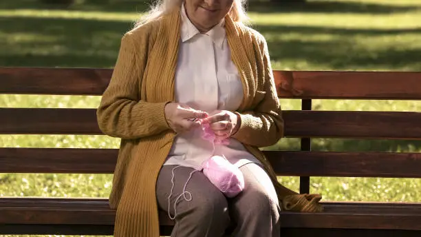 Old grandmother resting on bench in park, knitting for newborn grandchild family