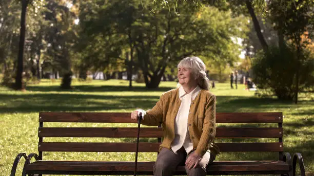 Old woman with walking stick enjoying sunny day in park, sitting on bench