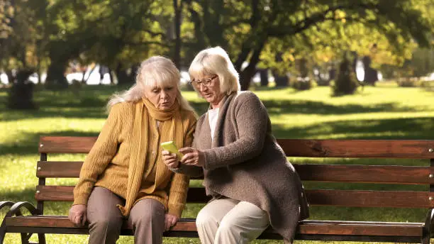 Old lady showing news feed on mobile phone to friend, sitting on bench in park