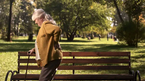 Senior lady slowly walking with stick along park suffering from back pain