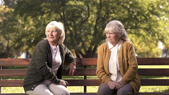 Two grumpy old ladies judging passerby people, sitting on bench in park, pension