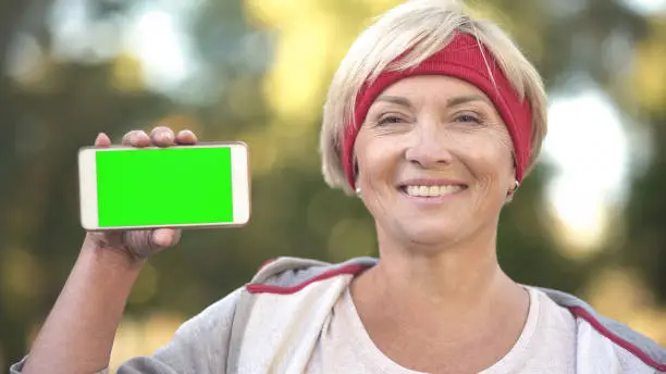 Smiling fit middle-aged woman showing green screen mobile phone, sports app