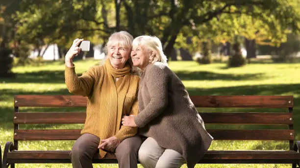 Two senior women taking selfie on cellphone, sitting on bench in park technology