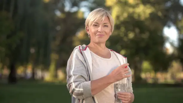 Pretty middle aged woman drinking water in park, keeping water balance, health