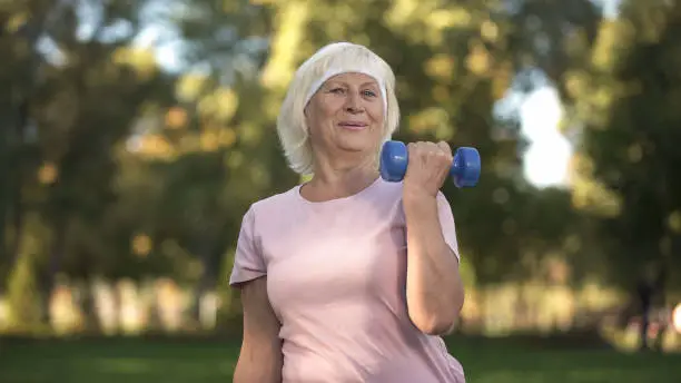 Smiling elder lady doing fitness with dumbbells at sunny day in park, lifestyle
