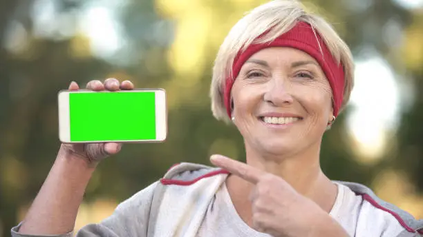 Sportive adult lady pointing at smartphone with green screen and smiling, app