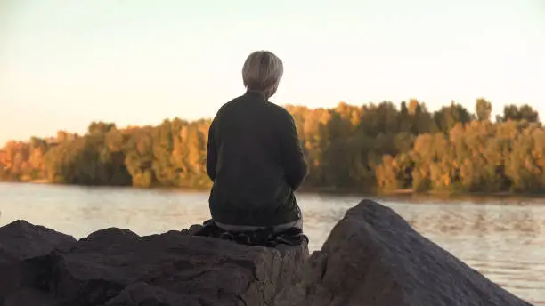 Photo of Lonely old woman sitting near lake and throwing stones solitude, back view