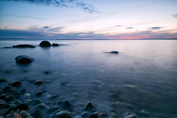 This evocative image offers a ground-level view of a pebble-strewn beach leading to the tranquil sea, with the soft blur of beachgoers in the distance. The foreground of smooth stones draws the eye, suggesting the tactile pleasure of a beachcombing wander, while the out-of-focus background invites contemplation of summer days by the water. Ideal for themes of travel, leisure, and the allure of the seaside.