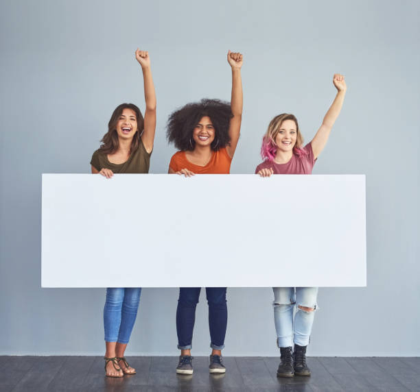 Here's to all the strong women out there Studio shot of a group of young women holding a blank placard and cheering against a gray background cheering group of people success looking at camera stock pictures, royalty-free photos & images