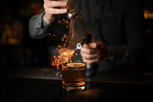 Bartender adding to a cocktail with ice cube and dry orange on it a cinnamon and flaming it with a burner on the bar counter in the dark.