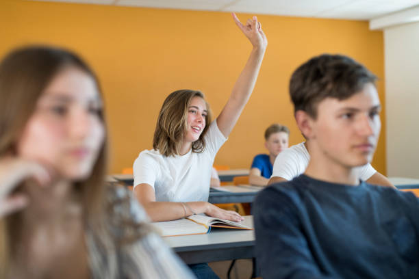 Girl raising hand to ask doubts in classroom Teenage girl raising hand to ask doubts in classroom. She is learning with friends in high school. Confident student is sitting at desk. hand raised classroom student high school student stock pictures, royalty-free photos & images