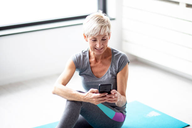istock Modern short blonde hair mature woman using mobile phone while relax after exercise in corner of room with window at background 1137734766