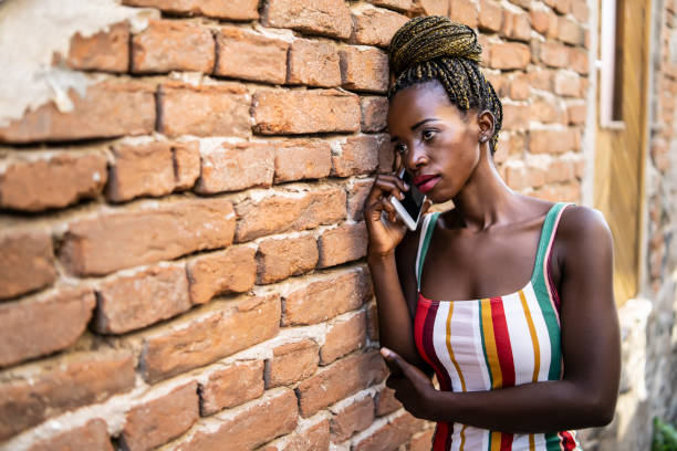 Sad african woman talking on smart phone, on street, leaning on brick wall.