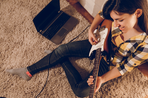 Young woman is using laptop to learn how to play electric guitar