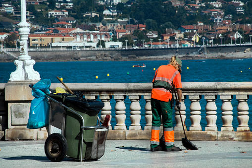 Gijon, Spain-August 31, 2018: Rear view of female street sweeper with cart, garbage can, broom and dustpan working on the sidewalk, waterfront by the beach, sea in the background, summer day in San Lorenzo beach in Gijón, Asturias, Spain..