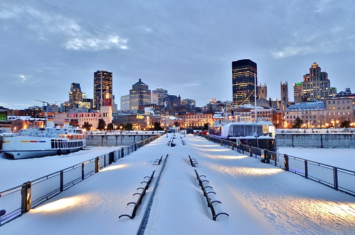 Snow-covered benches, pier, waterfront walkway, harbour, and downtown Montreal after sunset in winter - Montreal, Canada