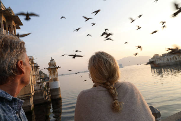 Couple look out to Floating Palace on lake at sunset They are at a ghat and pigeons fly overhead lake palace stock pictures, royalty-free photos & images