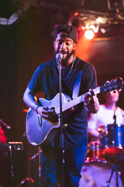 Photo of Black male guitarist singing and playing acoustic guitar on stage