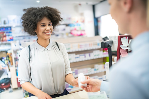 Young smiling African American woman receiving pill bottle over the counter from a female pharmacist.