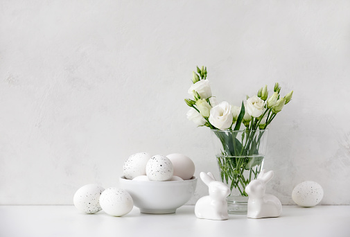 Easter greeting background, front view on white table decorated with  flowers in a vase, traditional eggs and ceramic rabbits, blank space for a text