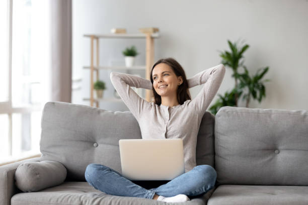 felice donna sorridente che si allunga, rilassante su un accogliente divano con laptop - women dependency business time foto e immagini stock