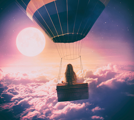 Dreamy photo manipulation of little girl flying with a hot air balloon over the clouds, looking at full moon.
