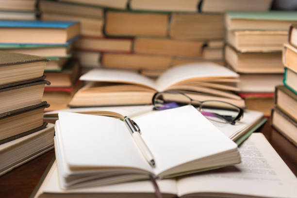 istock Education learning concept with open book and notebook on the table. Stack piles of books on reading desk and glasses in bookshelves background 1137832348