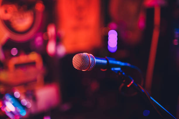 istock Microphone on a stand ready for live music performance or karaoke night with soft bokeh lights and people silhouettes in the background. Concept for musical singing event, having a good time. 1137845548