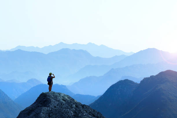 Female Hiker Taking Picture Of Sunset A female hiker stands on top of a rocky promontory as she uses her mobile phone to take a picture of the setting sun on the distant horizon. A series of mountain ridges seem to disappear into the distance as the haze obscures the details creating a graphic outline of the various layers of ridge lines. view from mountain top stock pictures, royalty-free photos & images