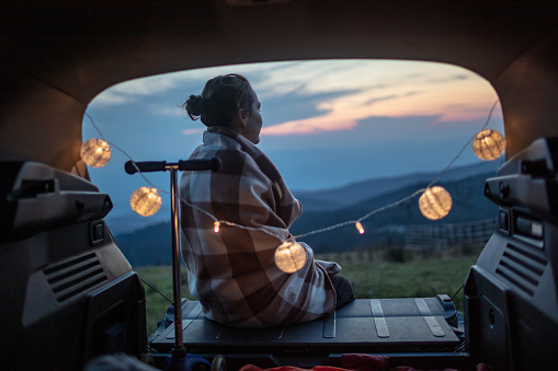 Beautiful woman curled in a blanket resting in a car trunk and looking at view