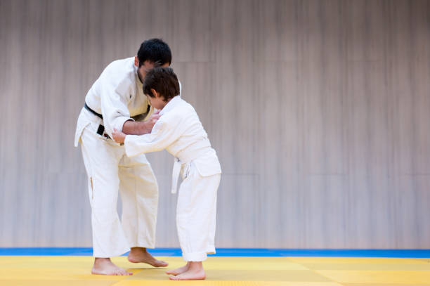 トレーナーと若い柔道家は道場で柔道のクラスに従事しています。トレーナーは子供に単一の戦闘、柔道、空手や合気道の方法と位置を教えます - child sport karate education ストックフォトと画像