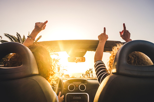 Couple of woman friends traveling and driving having a lot of fun dancing in the car with opened roof and summer vacation sunset ocean in front - concept of friendship together and nice lifestyle for independent girls