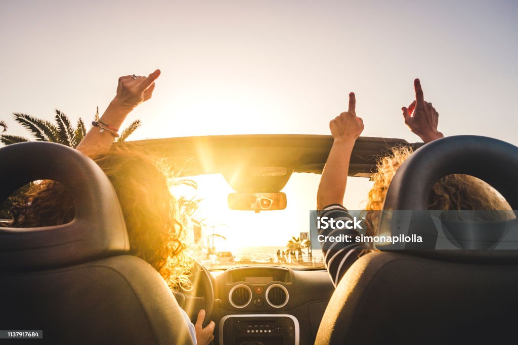 Couple of woman friends traveling and driving having a lot of fun dancing in the car with opened roof and summer vacation sunset ocean in front - concept of friendship together and nice lifestyle for independent girls Car Stock Photo