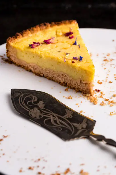 One piece of vegan lemon cheesecake, garnished with eatable petals, on a white plate with ornate cake server and some crumbs - close-up, vertical portrait orientation