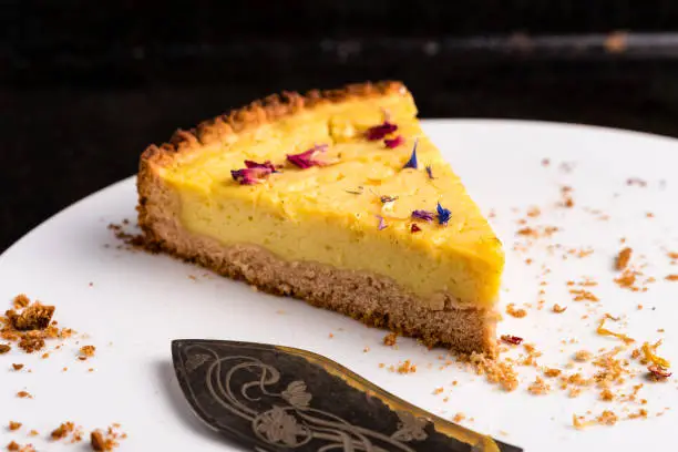 One piece of vegan lemon cheesecake, garnished with eatable petals, on a white plate with ornate cake server and some crumbs - close-up, horizontal landscape orientation