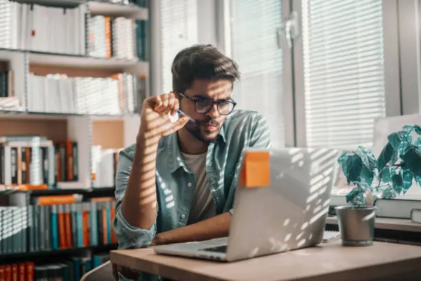 Photo of Serious mixed race male student with serious face sitting at the deck in library and using laptop for school project.