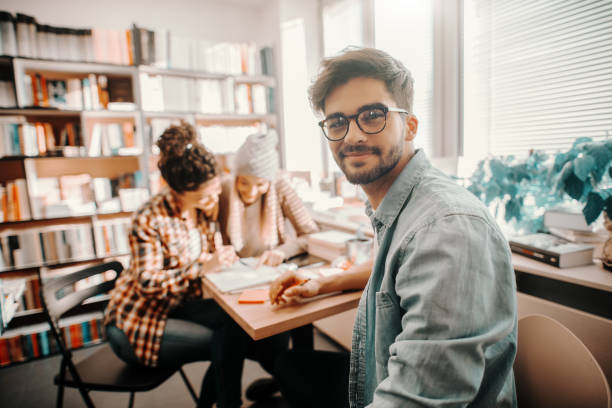 studente maschio di razza mista sorridente seduto alla scrivania, guardando la macchina fotografica e studiando per gli esami. sullo sfondo i suoi amici che studiano. interni della biblioteca. - hard life foto e immagini stock