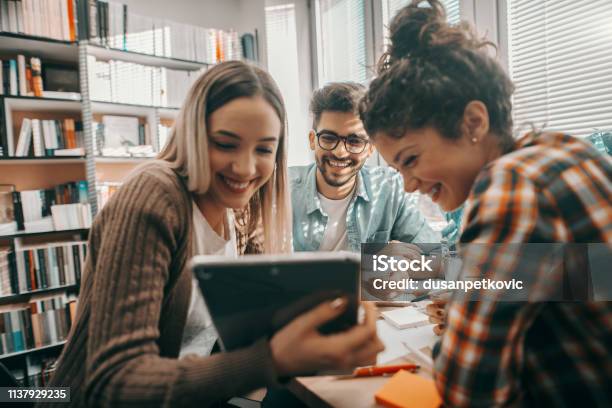 Three Happy Students Dressed Casual Using Tablet For School Project And Sitting At Desk In Library Stock Photo - Download Image Now