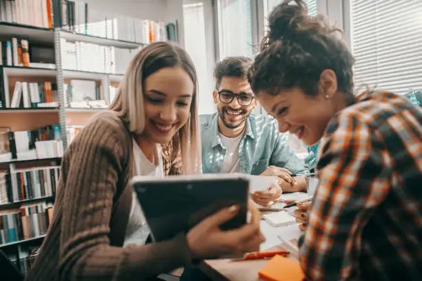 Photo of Three happy students dressed casual using tablet for school project and sitting at desk in library.